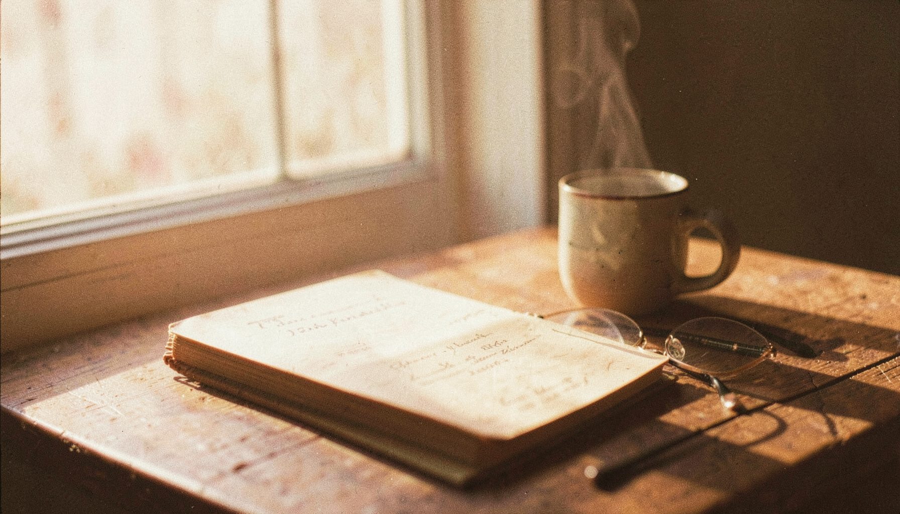 A warm, softly lit still life of a well-worn journal, a steaming ceramic mug, and a pair of reading glasses resting on a wooden table near a window, with gentle morning light casting long shadows. Analog film photograph with visible grain, cream and amber tones.